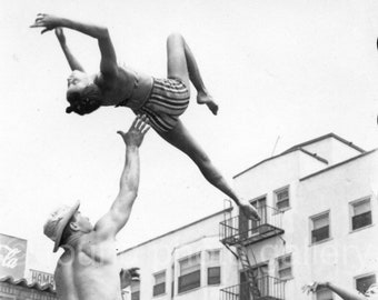 May include: A black and white photo of a man holding a woman in the air. The woman is wearing a striped swimsuit and is in a handstand position. The man is wearing a hat and swim trunks. There are people in the background watching the performance.