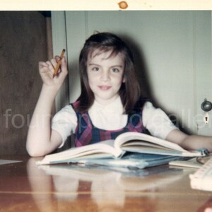 May include: A young girl with brown hair sits at a wooden table, holding a pencil in her right hand. She is looking at the camera and smiling. She has a book open in front of her.