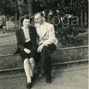 May include: A black and white photo of a couple sitting on a bench outdoors. The woman is wearing a dark jacket and a skirt. The man is wearing a suit jacket and pants. They are both smiling.