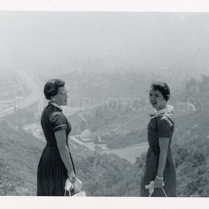 Puede incluir: Dos mujeres con vestidos se encuentran en la cima de una colina con vistas a una ciudad. La mujer de la izquierda lleva un vestido oscuro y gafas. La mujer de la derecha lleva un vestido claro con un cuello blanco y sonríe. La ciudad está en el fondo y está brumosa.