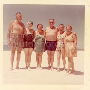 May include: A group of six people, including three women and three men, stand on a sandy beach. The women are wearing swimsuits, and the men are wearing swim trunks. The people are smiling and looking at the camera.
