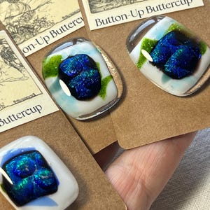 May include: Three square glass buttons attached to brown cardboard display cards labeled Button-Up Buttercup. Each button features a white base with a central iridescent dark blue and green textured glass inlay and a smooth, glossy finish.