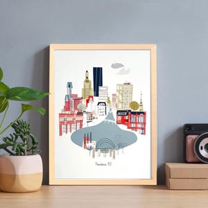 Pode incluir: Uma impressão artística emoldurada com uma ilustração colorida de um horizonte urbano com edifícios em vermelho, azul e dourado. A impressão é exibida em uma superfície de madeira ao lado de plantas em vasos e uma câmera.