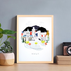 May include: A framed art print depicting Salt Lake City, Utah, with a colourful illustration of the city skyline and mountains. The print is in a light wood frame, and the text "Salt Lake City, UT" is at the bottom.