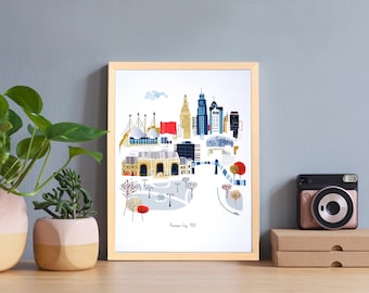 Könnte beinhalten: Ein gerahmter Kunstdruck mit einer farbenfrohen Illustration einer Stadtlandschaft, mit Gebäuden in Blau, Gelb und Rot. Der Druck befindet sich in einem hellen Holzrahmen und wird auf einer Holzoberfläche präsentiert. Der Text "Kansas City, MO" ist sichtbar.