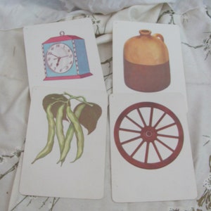 May include: Four vintage flashcards with illustrations of a clock, a jug, green beans, and a wagon wheel. The clock has a white face with black numbers and hands. The jug is brown and has a white spout. The green beans are green and have brown stems. The wagon wheel is red and has a black center.