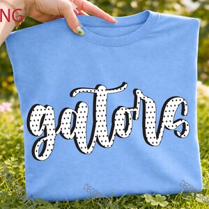 May include: A light blue t-shirt featuring the word "gators" printed in a bold, cursive font. The lettering is white with black polka dots and a thick black drop shadow, creating a layered effect against the solid light blue fabric background.