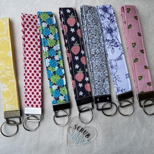 May include: A collection of seven fabric wrist keychains with various patterns. Designs include yellow floral, red polka dots, blue and green flowers, strawberries, small floral, blue floral, and pink stripes with strawberries. Each keychain has a metal ring.