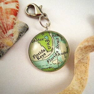 May include: A round silver charm featuring a map of the Florida Keys and surrounding areas in shades of green and blue. The charm is attached to a silver clasp. The charm is a travel-themed accessory.