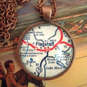 May include: A copper-colored pendant necklace featuring a round charm with a map of Flagstaff, Arizona. The map includes roads, landmarks, and the text "Flagstaff." The pendant hangs from a matching chain.