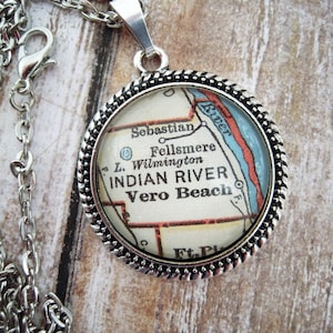 May include: Silver-toned pendant necklace featuring a circular charm with a map of Indian River, Vero Beach, and surrounding areas. The map is encased in a decorative silver frame and hangs from a delicate chain. The pendant's design includes the text "INDIAN RIVER" and "Vero Beach".