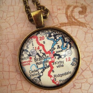 May include: A round pendant necklace with a map design. The pendant has a brass-colored frame and chain. The map features red, blue, and black lines, with the word "Branson" visible. The background is a textured, light pink surface.
