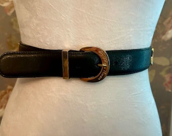 May include: Black leather belt with a gold-tone buckle. The buckle has an engraved design and the words "House of Versace". The belt is displayed on a mannequin.