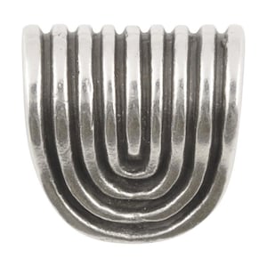 May include: A silver-toned metal decorative object with a half-circle shape. The design features a series of vertical and curved lines, creating a textured, ridged surface. The object has a vintage or antique appearance.