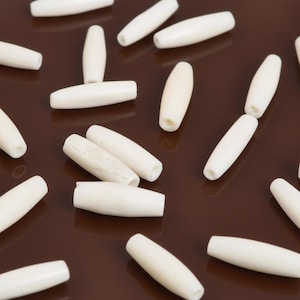 May include: Ivory-colored, elongated beads scattered on a brown surface. The beads have a smooth, cylindrical shape with rounded ends and a hole running through the center. These beads are suitable for jewelry making and crafting projects.