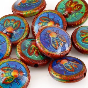 May include: A collection of round, red, iridescent glass beads with a raised image of a fly in the center. The flies are painted in shades of orange, yellow, and red, with green and blue accents. The beads have a decorative border around the edge.