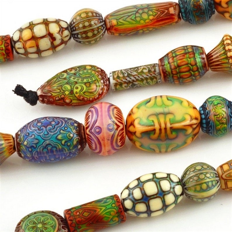 May include: A close-up of a string of decorative beads in various shapes and colours. The beads have intricate patterns in orange, green, blue, and cream. Some are oval, others cylindrical, and some are teardrop-shaped, showcasing detailed craftsmanship.