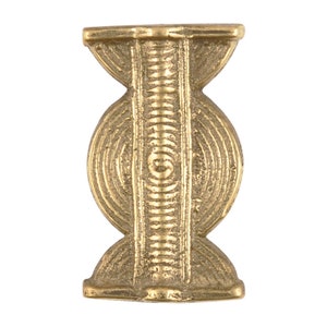 May include: A brass-colored, symmetrical decorative object. The design features a central vertical column with a series of horizontal lines, flanked by two circular shapes with concentric circles. The top and bottom have a flared, curved shape.