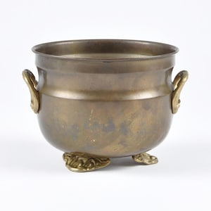 May include: A round brass pot with a smooth, weathered finish, featuring two small decorative side handles and three ornate, sculpted feet. The metal surface displays a muted, brownish-gold tone with subtle variations in color and texture.