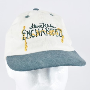 May include: An off-white baseball cap with a dark teal brim. The front features embroidered text that reads Stevie Nicks in black script above the word ENCHANTED in dark green block letters with decorative gold vine-like accents.