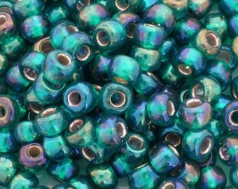 May include: A close-up shot of a pile of iridescent teal glass beads. The beads have a metallic sheen with hints of purple and gold. Each bead is cylindrical with a large hole running through the center, perfect for stringing.