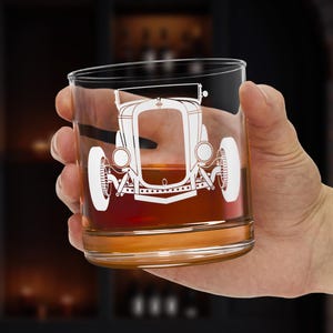 Könnte beinhalten: Ein klares Glas mit einem weißen Oldtimer-Design. Das Glas ist mit einer braunen Flüssigkeit gefüllt. Das Glas wird von einer Person in der Hand gehalten.