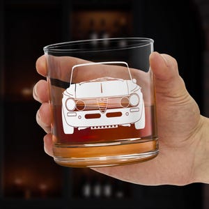 May include: A clear, lowball whiskey glass with a white etched design of a vintage car. The glass is filled with a golden-brown liquid. The design features the car's front, including headlights, grille, and bumper.