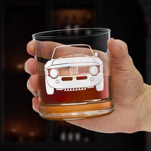 以下が含まれることがあります： 前面に白い車のグラフィックがエッチングされた、透明で丸いウイスキーグラス。グラスには茶色の液体が入っており、人の手で持たれています。背景は暗いバーの設定です。