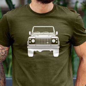 Puede incluir: Camiseta verde oliva con un contorno blanco de un vehículo todoterreno clásico. El gráfico está centrado en el pecho. La camiseta tiene mangas cortas y cuello redondo.