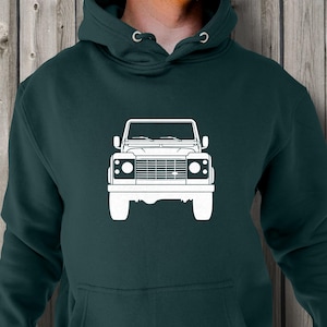 Puede incluir: Sudadera con capucha verde oscuro con un gráfico blanco de la parte delantera de un vehículo. La sudadera tiene una capucha con cordones y un bolsillo delantero. El gráfico del vehículo está centrado.