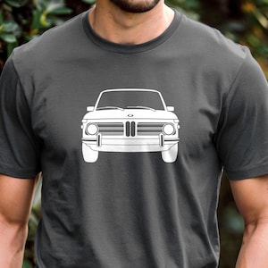 May include: Dark grey t-shirt with a white outline of a classic car's front. The car design includes headlights, a grille, and a bumper. The t-shirt is made of a soft material and is suitable for casual wear.