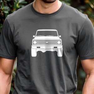 May include: Dark gray t-shirt featuring a white outline of a classic pickup truck. The graphic shows the front view of the vehicle, including the headlights, grille, and bumper. The shirt is a crew neck style.