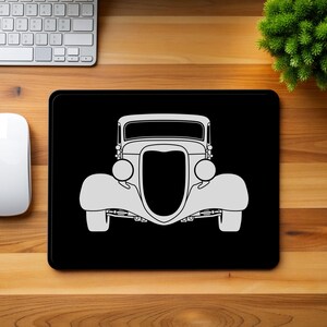 Puede incluir: Alfombrilla de ratón rectangular negra con una ilustración de coche antiguo blanco. El coche es un modelo clásico con faros redondos y una parrilla prominente. La alfombrilla está sobre un escritorio de madera con un teclado, un ratón y una pequeña planta verde.