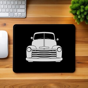 Peut inclure: Tapis de souris rectangulaire noir avec un dessin au trait blanc d'un pick-up vintage. Le tapis de souris est posé sur un bureau en bois, avec une souris blanche, un clavier et une petite plante verte.