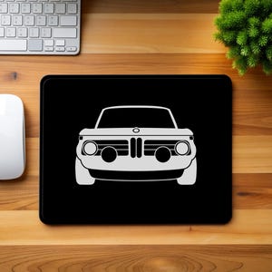 Puede incluir: Alfombrilla de ratón rectangular negra con un gráfico blanco de la vista frontal de un coche clásico. La alfombrilla está sobre un escritorio de madera, junto a un ratón y un teclado blancos. Una pequeña planta verde está en la esquina superior derecha.