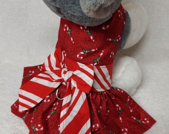 Puede incluir: Un vestido rojo para perro con un estampado de bastones de caramelo y un gran lazo a rayas rojas y blancas. El vestido tiene una falda con volantes y un pequeño anillo de metal adjunto al lazo.