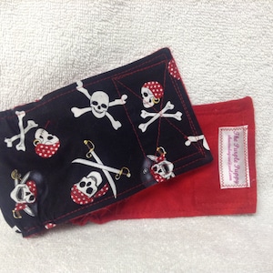 May include: A black and red fabric baby diaper cover with a pirate skull and crossbones pattern. The cover has a red fabric lining and a white label that says "The Purple Pippy".