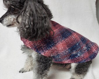 May include: A gray poodle wearing a red and blue plaid dog coat. The dog is sitting on a white surface. The coat is made of a soft, woven fabric and covers the dog's back and chest.