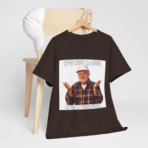 Può includere: Una t-shirt a maniche corte marrone scuro con una stampa grafica di una persona che indossa un casco bianco e una camicia a quadri con le mani alzate. Il testo sopra e sotto l'immagine recita IT'S NOT A BUG IT'S A FEATURE in lettere bianche.