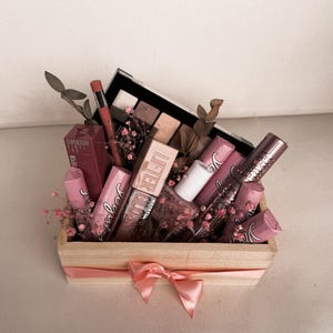 Könnte beinhalten: Eine Holzkiste gefüllt mit Make-up-Produkten, darunter Lippenstifte, Lipgloss und eine Lidschattenpalette. Die Produkte sind in Rosa- und Brauntönen gehalten, und die Kiste ist mit einem rosa Band und Trockenblumen verziert. Es ist ein Geschenkset.