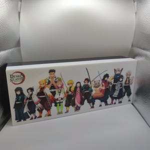 May include: A rectangular box with a white surface featuring a colorful illustration of characters from "Demon Slayer." The box has a black top and the "Demon Slayer" logo is visible. The characters are in various poses, with swords and unique outfits.