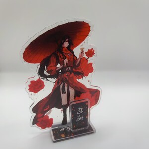Peut inclure: Un présentoir en acrylique transparent représentant un personnage en tenue rouge, tenant un parapluie rouge et une petite fleur. Le présentoir a une base avec du texte et un motif floral. Le personnage a de longs cheveux noirs et est entouré d'accents floraux rouges.