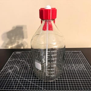 May include: A clear glass laboratory bottle with a red plastic screw cap and a white pouring ring. The bottle features white printed graduation markings ranging from 400 to 1800 ml, with a 2000 ml capacity label and a white rectangular marking area.