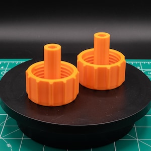 May include: Two orange, 3D-printed knobs with a cylindrical post and a ridged outer ring. The knobs are on a black, circular platform against a green cutting mat background. The knobs are likely for a mechanical or electronic device.