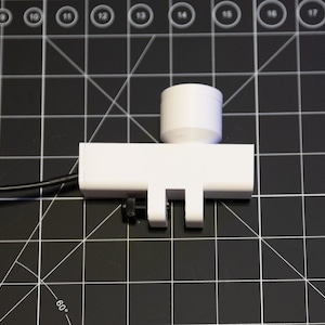 May include: A white, rectangular device with a cylindrical top and two downward-facing prongs. A black wire extends from the side. The device is on a black surface with a grid pattern.