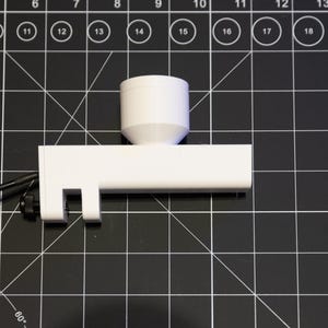 May include: A white 3D-printed object with a cylindrical top and a rectangular base, connected by a conical section. A black cable with a connector is attached to the base. The object is on a black grid surface.