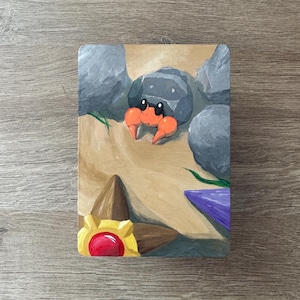 May include: A rectangular painting featuring a cartoon crab with orange claws and a gray shell, surrounded by gray rocks and sand. A yellow and red jewel is in the lower left corner, with a purple shape on the right.
