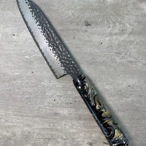 May include: A black and gold patterned handle kitchen knife with a hammered finish blade.