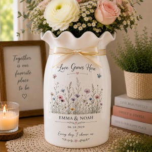 May include: A white ceramic vase with a ruffled rim, decorated with a floral meadow illustration, a beige ribbon, and the text Love Grows Here, Emma & Noah, 06.18.2019, and Every day, I choose us. The vase holds a bouquet of white and pink flowers.
