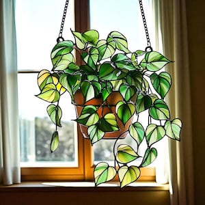 May include: Stained glass hanging planter with a trailing vine design. The leaves are shades of green and yellow, and the planter is a terracotta color. The piece hangs from a black chain in front of a window.